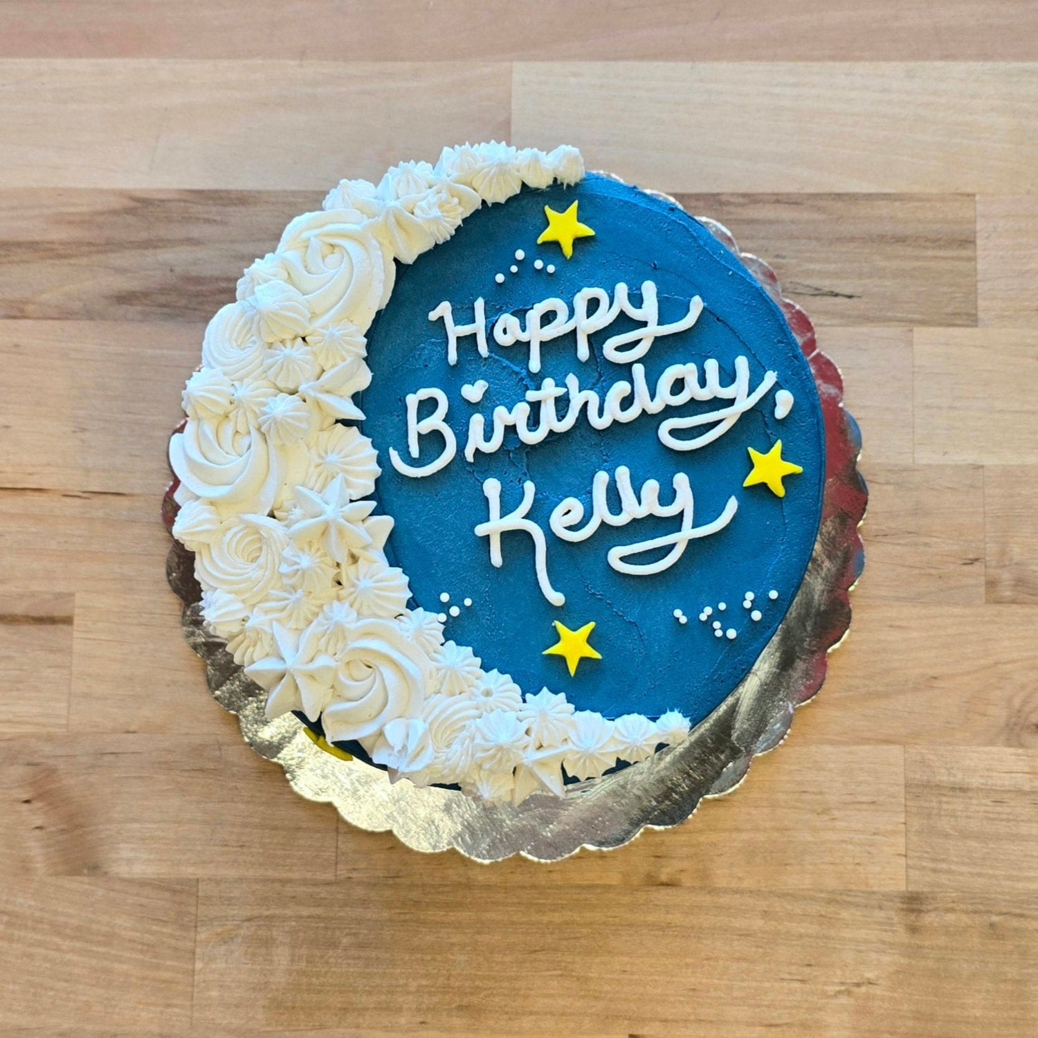 A pre-decorated To the Moon cake with dark blue frosting, a white frosting crescent moon, yellow fondant stars, and sprinkle constellations.