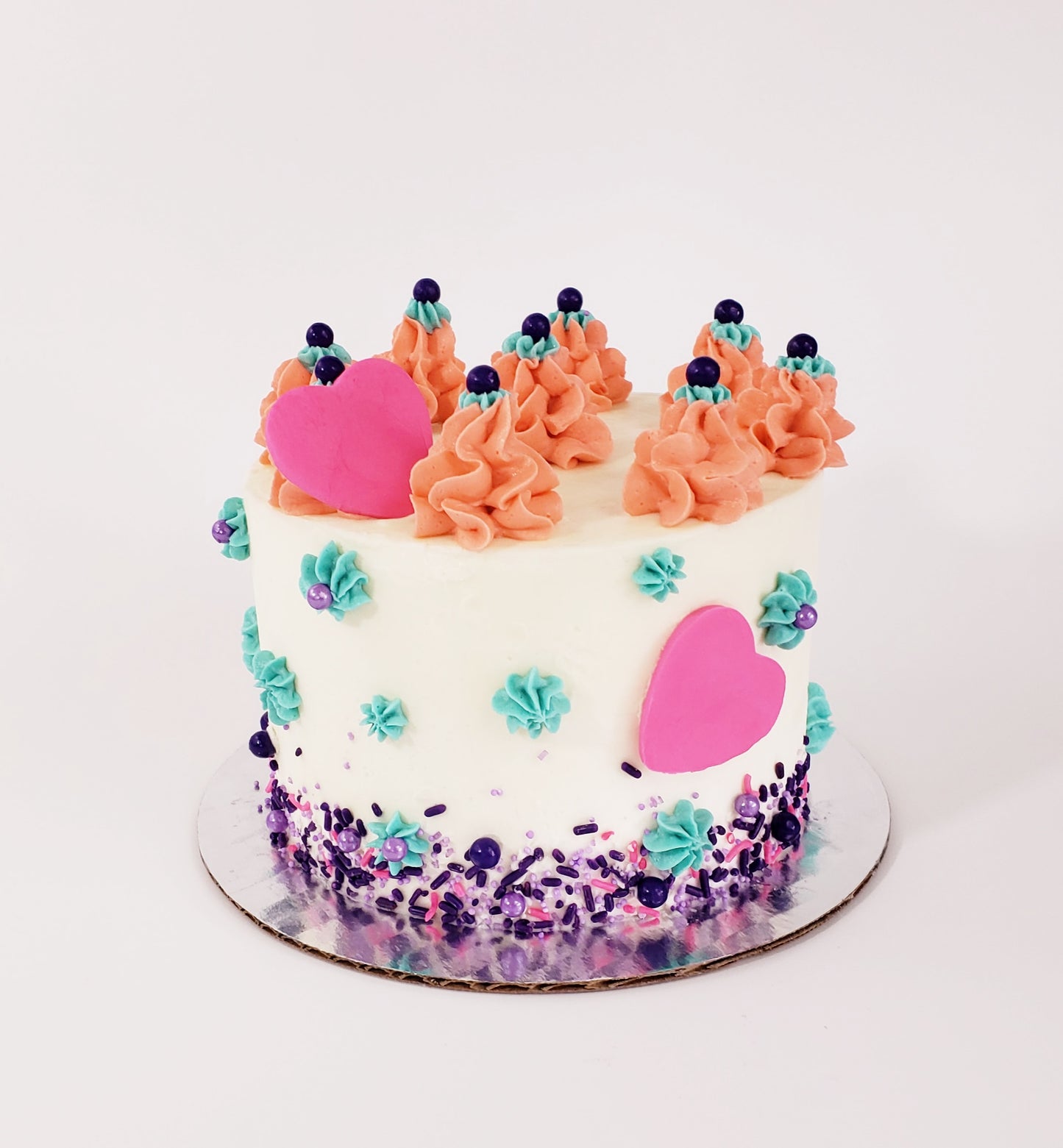 A white frosted cake with coral colored frosting mounds on top, teal frosting stars, pink hearts made of fondant, and pink and purple sprinkles around the bottom; a pre-decorated cake available for pick up at Cake Hoopla in Tigard, Oregon, in the Portland area.