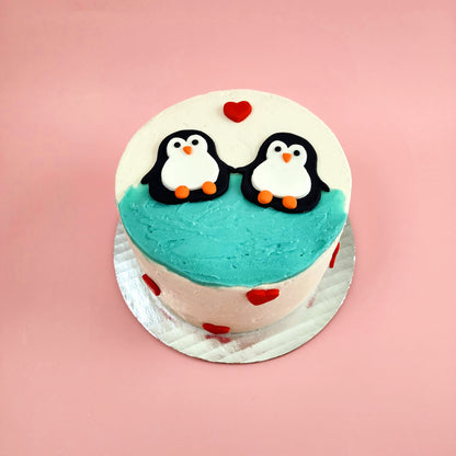 A love-themed white cake with teal blue frosting and two black, white, and orange penguins jumping into the water and red fondant hearts dotting the cake; a pre-decorated cake shipped to the continental US.