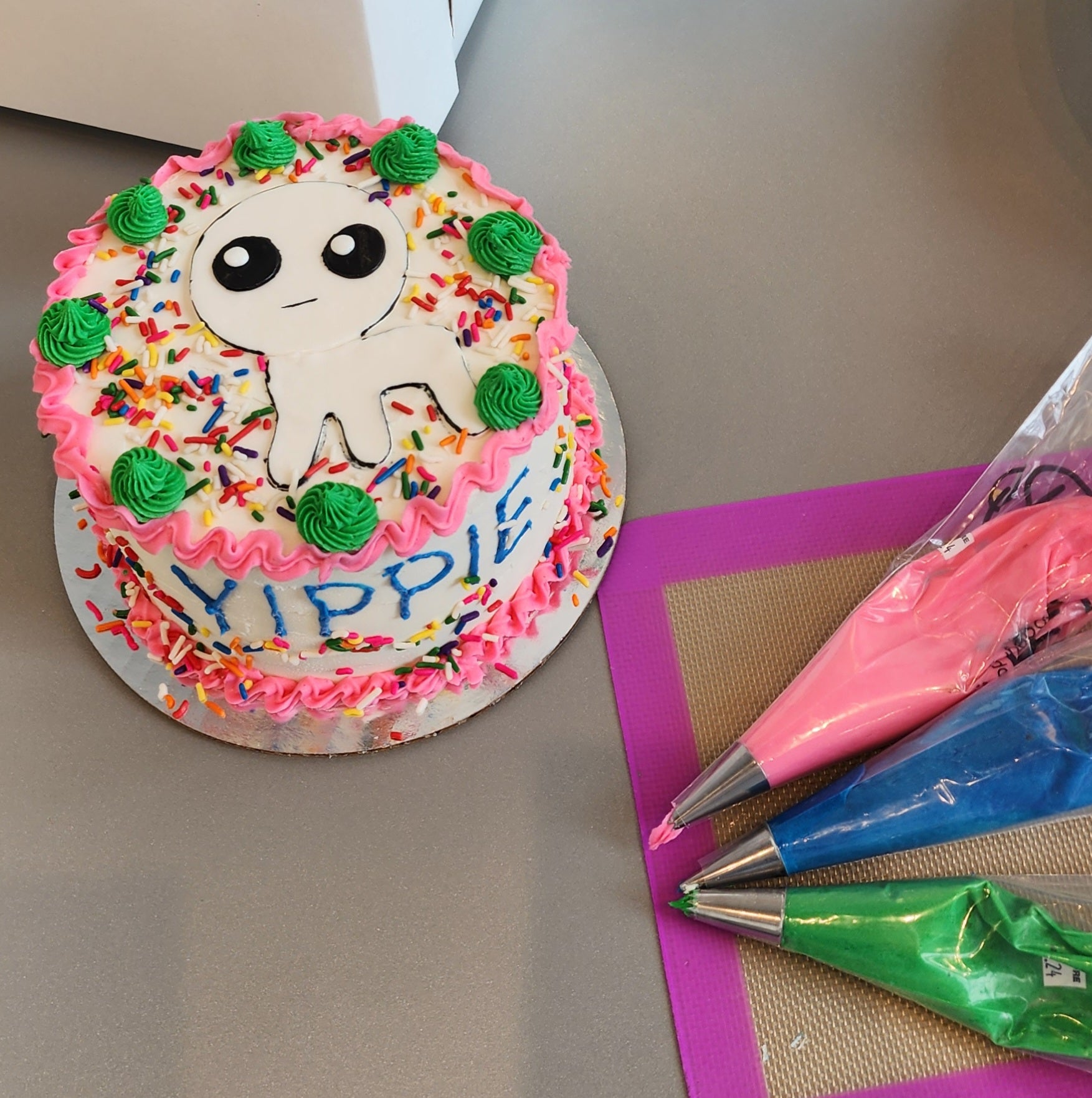 At the Cake Hoopla DIY cake/cupcake-decorating studio, a customer decorated a custom cake with a cute little creature on top, pink and green frosting borders, and YIPPIE written in blue frosting across the front.