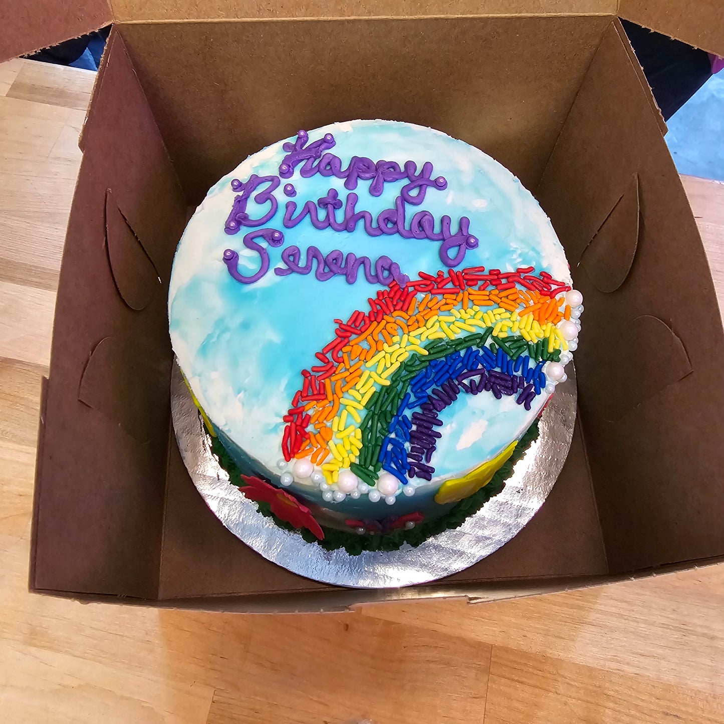 A birthday cake frosted with white and blue frosting, pink fondant flowers, and rainbow sprinkles from a Cake Hoopla Create Your Own Cake Kit available during open studio time, as a to-go kit, or as a shipped kit from Cake Hoopla in Tigard, Oregon near Portland.