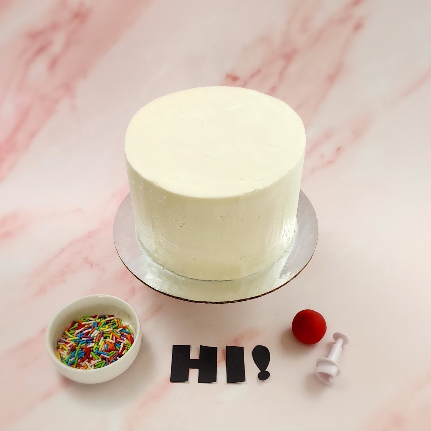 Components of a DIY cake-decorating kit "Just Hi!" with white frosted cake, rainbow sprinkles, letter cutouts, red fondant, and a heart cutter, available during open studio time or as a to-go kit from Cake Hoopla in Tigard, Oregon near Portland.