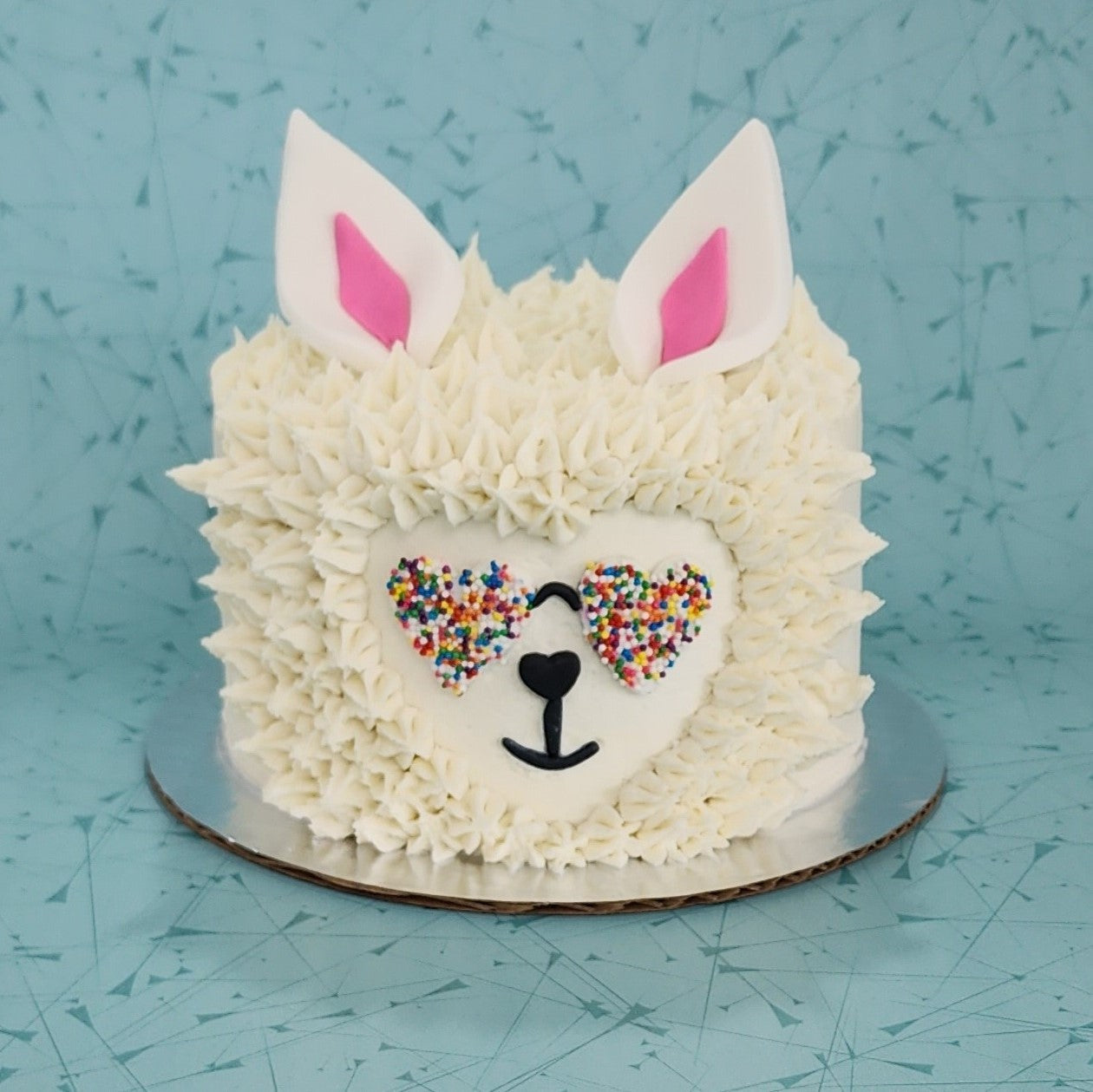 A llama-themed white cake that looks like a llama head with white frosting fur, heart rainbow sunglasses, and pink and white ears; a DIY cake kit available to decorate during open studio time at Cake Hoopla in Tigard, Oregon, in the Portland area.