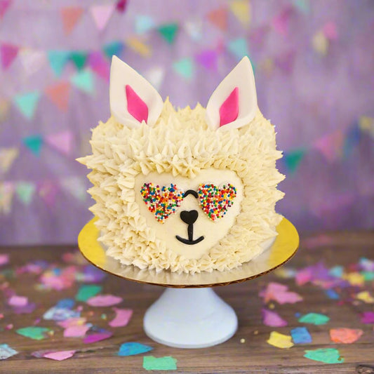 Cute llama cake with white frosting fur, heart-shaped rainbow-sprinkled sunglasses, and white ears with pink centers.