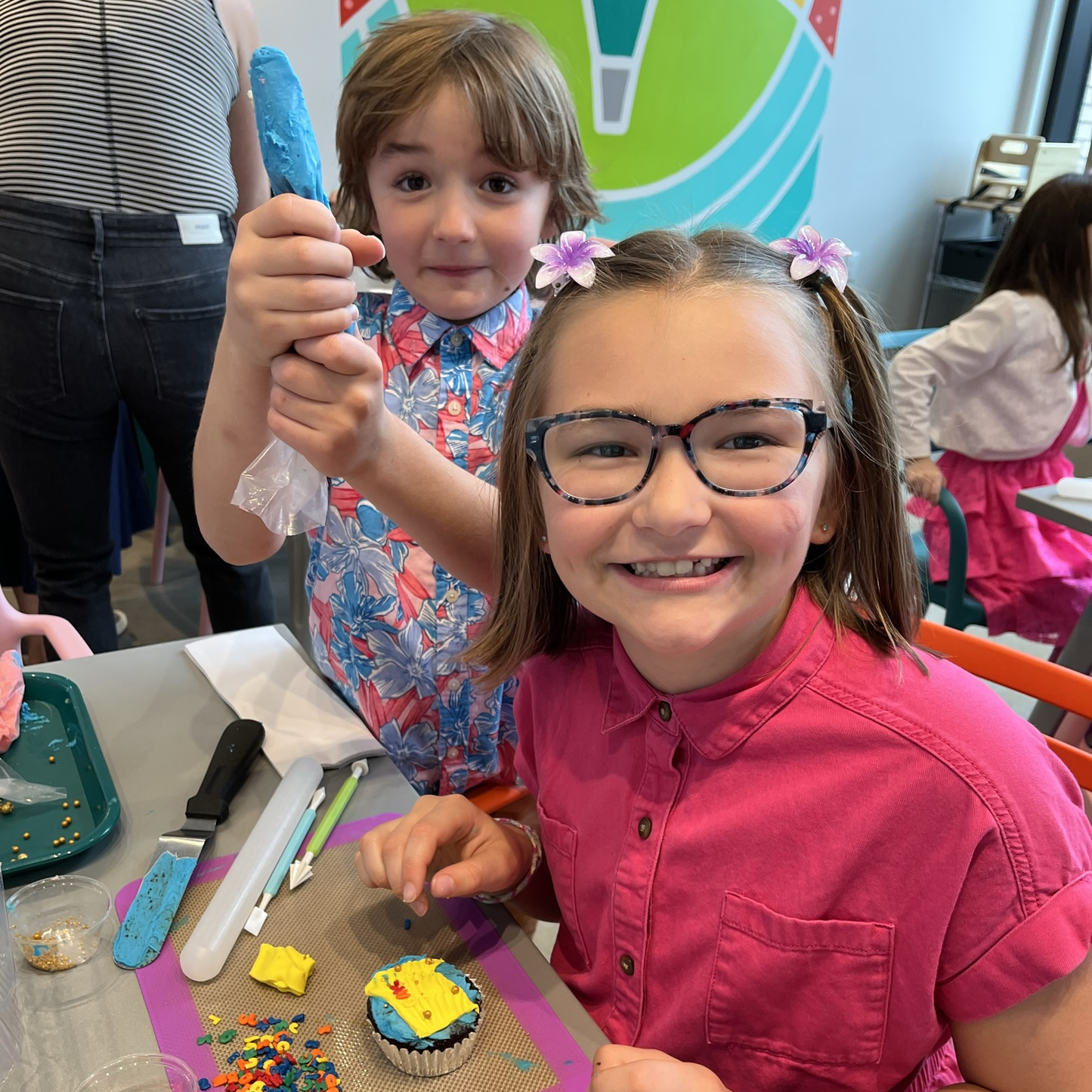 two small children, a boy and a girl, are decorating cupakes. The boy holds up a piping bag of blue frosting in a pose of triumph