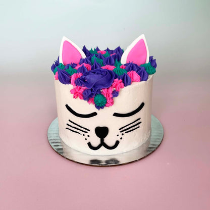 A cat-themed white cake made to look like a cat face with black facial details, purple, teal, and pink frosting hair, and pink and white ears; a pre-decorated cake shipped to the continental US.