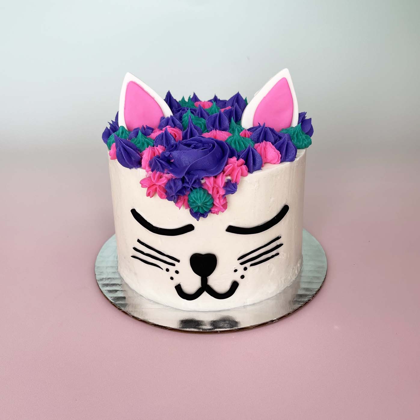 A cat-themed white cake made to look like a cat face with black facial details, purple, teal, and pink frosting hair, and pink and white ears; a DIY cake kit available to decorate during open studio time at Cake Hoopla in Tigard, Oregon, in the Portland area.