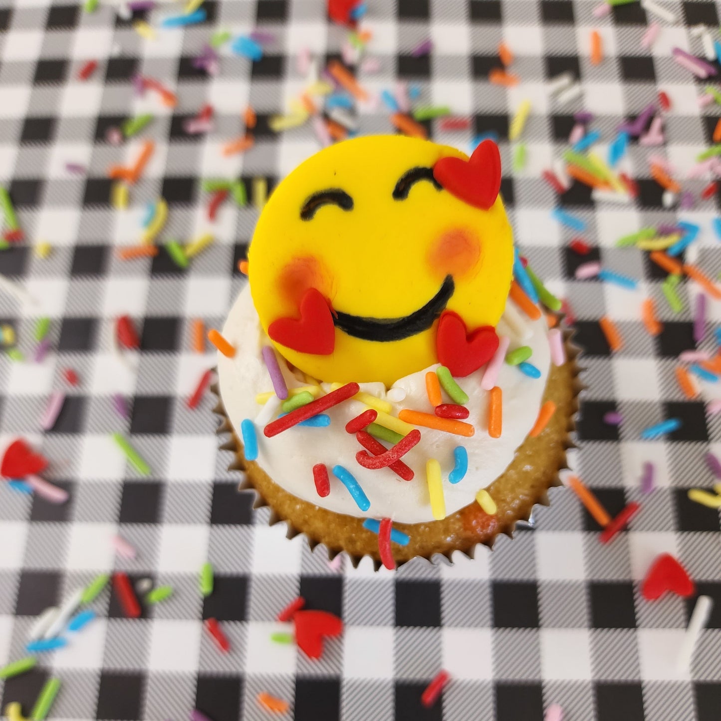 Emoji cupcakes frosted white with rainbow sprinkles and yellow smiling emoji faces with pink cheeks and three floating hearts; a DIY cupcake kit available to decorate during open studio time at Cake Hoopla in Tigard, Oregon, in the Portland area.