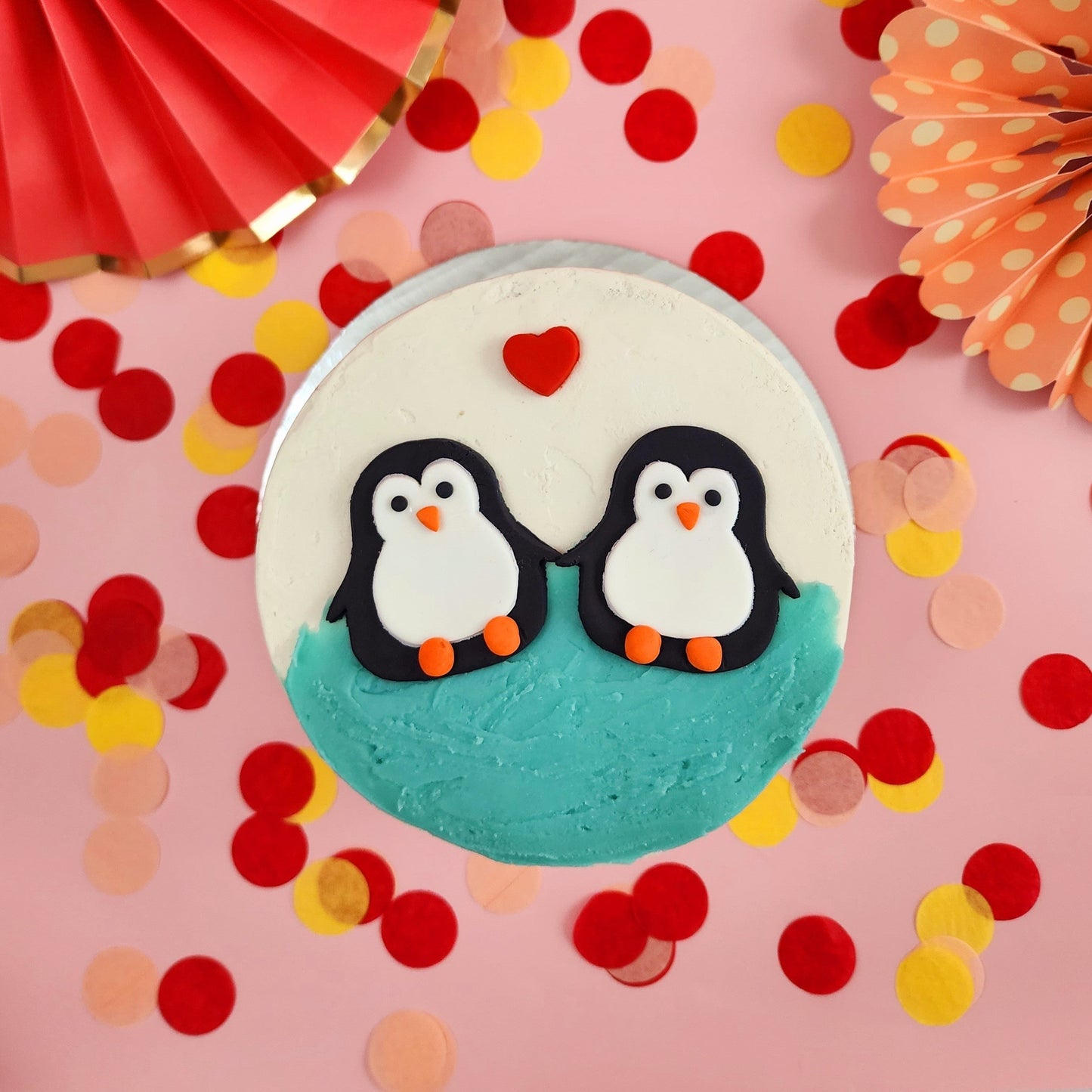 A love-themed white cake with teal blue frosting and two black, white, and orange penguins jumping into the water and red fondant hearts dotting the cake; a DIY cake kit available to decorate during open studio time at Cake Hoopla in Tigard, Oregon, in the Portland area.