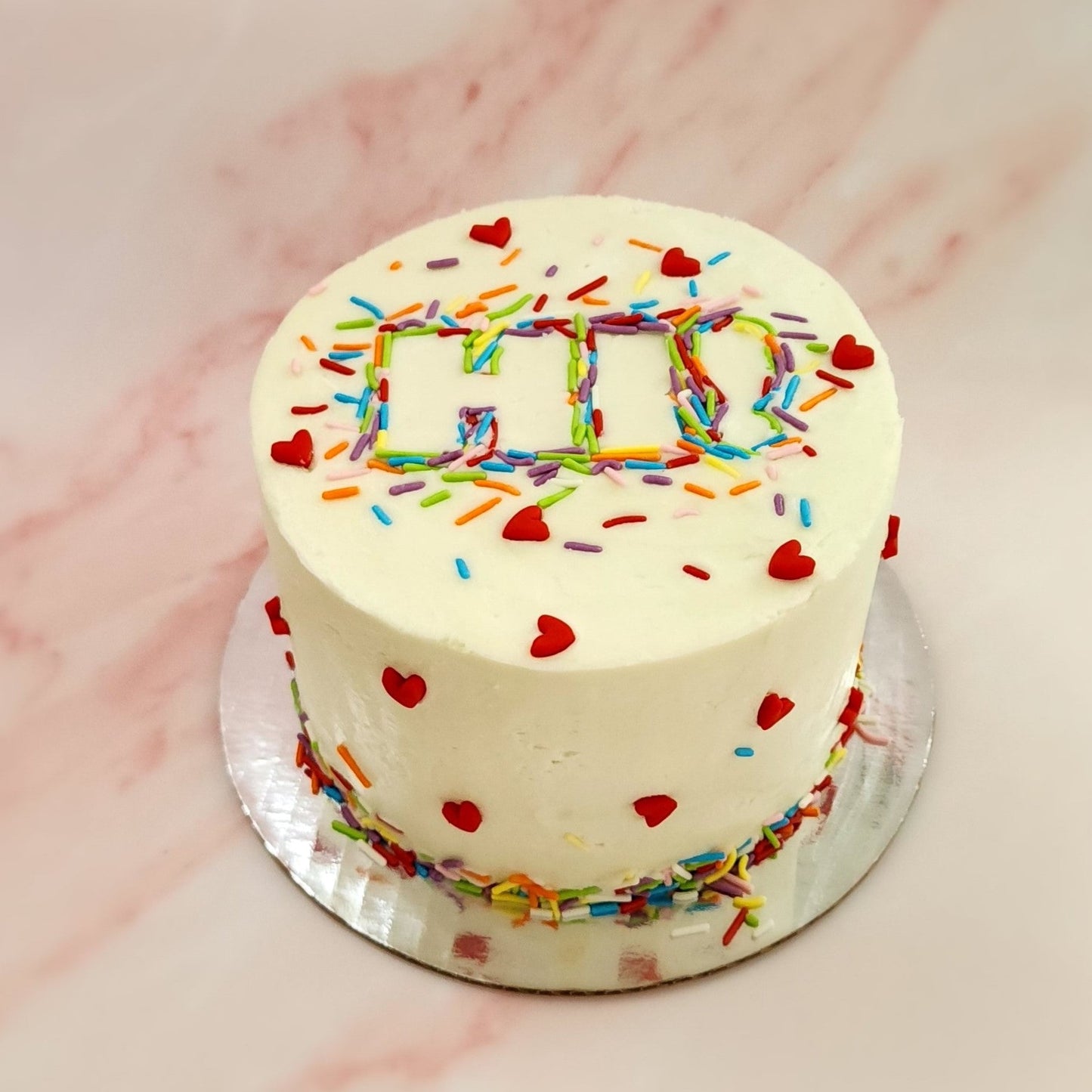 A fun white cake with the word "Hi!" spelled out of sprinkles and red fondant hearts dotting the cake; a DIY cake kit available to decorate during open studio time at Cake Hoopla in Tigard, Oregon, in the Portland area.