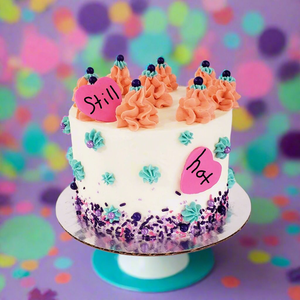 A bright, springtime array of colors on a white frosted cake. Teal frosting stars dot the sides of the cake, mounds of peach-colored frosting circle the top of the cake, and a purple sprinkle mix lines the bottom of the cake. One bright pink fondant heart rests against a frosting mound on top of the cake, and one is pressed into the frosting on the side of the cake. Available for shipping to the continental US.