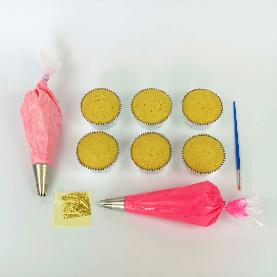 The components of the La Fleur DIY cupcake-decorating kit, including gold leaf, a paintbrush, cupcakes, light pink and dark pink frosting in piping bags, each with a different silver piping tip. Available during open studio time or as to-go kit at Cake Hoopla in Tigard, Oregon near Portland.