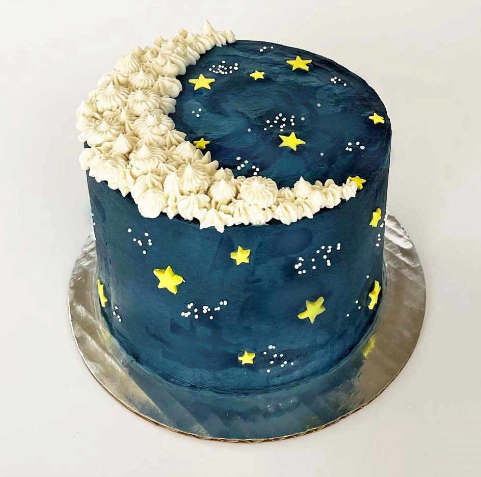 A moon-themed dark blue cake with a white crescent moon piped on the top with yellow fondant stars and white sprinkle galaxies dotting the cake; a DIY cake kit available to decorate during open studio time at Cake Hoopla in Tigard, Oregon, in the Portland area.