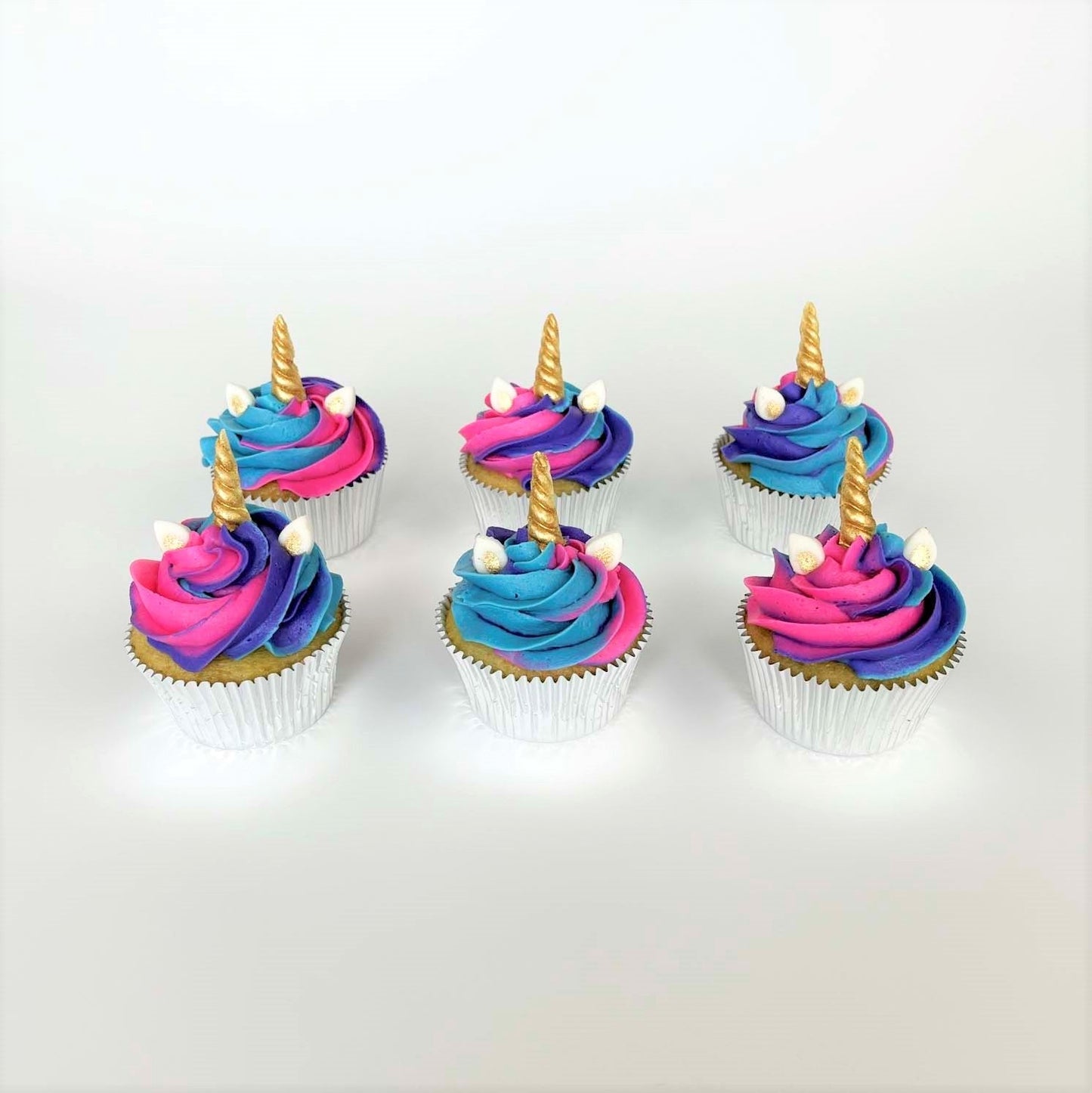 Unicorn-themed cupcakes frosted in blue, purple, and pink swirled frosted, white and gold ears, and gold unicorn horns; a DIY cupcake kit available to decorate during open studio time at Cake Hoopla in Tigard, Oregon, in the Portland area.