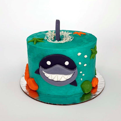 Shark-themed cake frosted in teal with green and orange shells and a shark face and tail coming out of the cake; a pre-decorated cake shipped to the continental US.