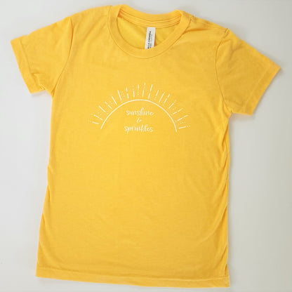 A soft sunflower-yellow short-sleeved t-shirt has white screen printing of the words “sunshine & sprinkles” written in a whimsical, almost cursive font across the center chest. The words are framed by an abstract half sun with rays of sprinkley sunshine shooting out.