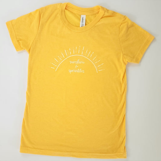 A soft sunflower-yellow short-sleeved t-shirt has white screen printing of the words “sunshine & sprinkles” written in a whimsical, almost cursive font across the center chest. The words are framed by an abstract half sun with rays of sprinkley sunshine shooting out.