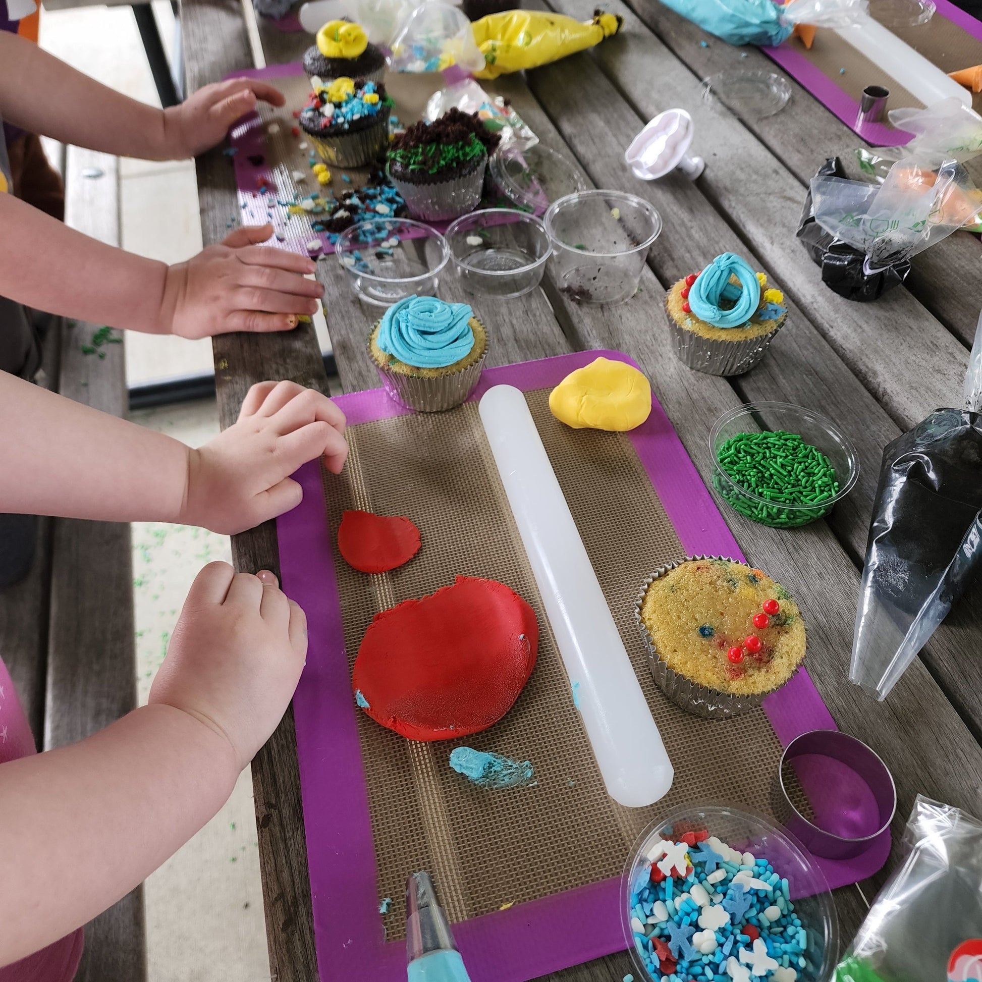 Little hands decorating cupcakes at a custom cupcake-decorating party in the Portland area. A picnic table is topped with a smattering of cupcake-decorating ingredients and tools, like fondant, rollers, cutters, frosting, sprinkles, and edible dirt.