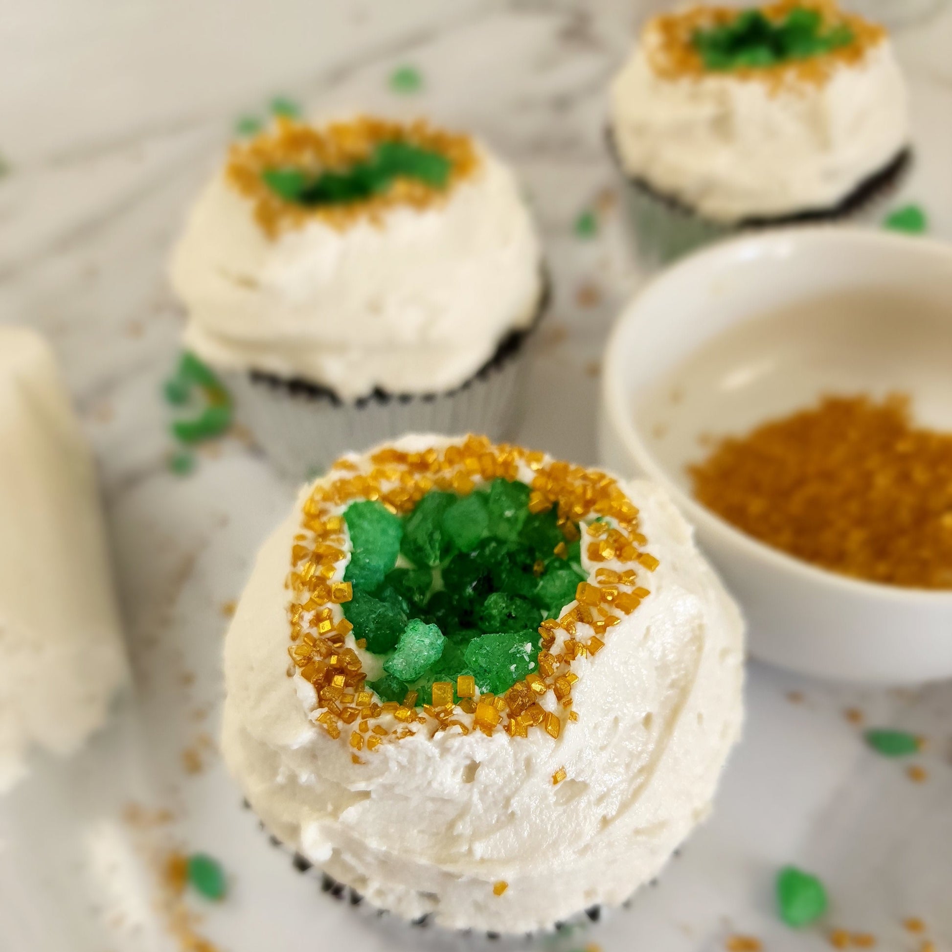 A closeup of one of the finished geode cupcakes from the You’re a Gem cupcake kit. White vanilla buttercream frosting glistens around gold sparkly sprinkles around green rock candy that forms a geode shape in the middle of the cupcake. 