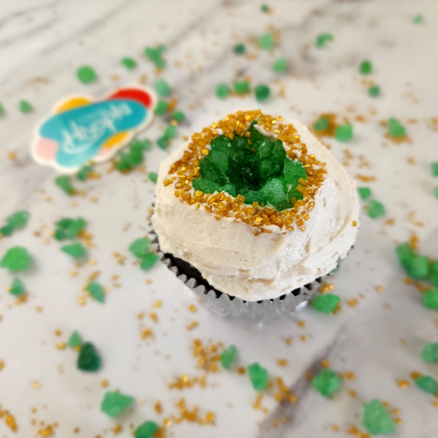 A closeup of one of the finished geode cupcakes from the You’re a Gem cupcake kit. White vanilla buttercream frosting glistens around gold sparkly sprinkles around green rock candy that forms a geode shape in the middle of the cupcake.