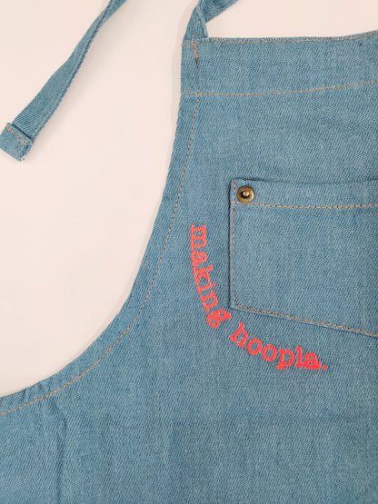 A closeup view of the corner of the breast pocket outlined with neon pink embroidery reading "making hoopla." The font has a typewritten look and contrasts nicely against the blue of the apron. Kids will stay clean and stylish in this apron.