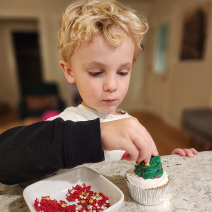 Adorable small child does DIY cupcake decorating, topping a frosting Christmas tree with sprinkles as ornaments.