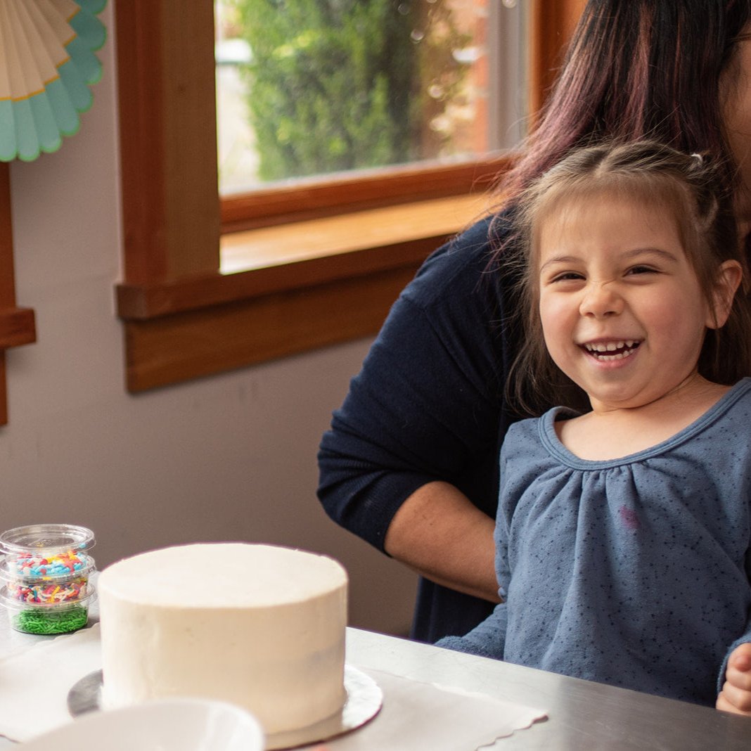 Happy little girl at a custom DIY cake decorating party in the Portland area, smiling big with an undecorated cake, sprinkles, and fondant in front of her. Ready to begin her DIY cake decorating, as her mom sits smiling behind her.