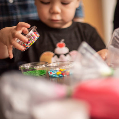 A close-up picture of a little boy at a custom DIY cake party in the Portland area. The boy holds a container of sprinkles. Other cups of sprinkles and piping bags of colorful frosting rest on the table in front of him.