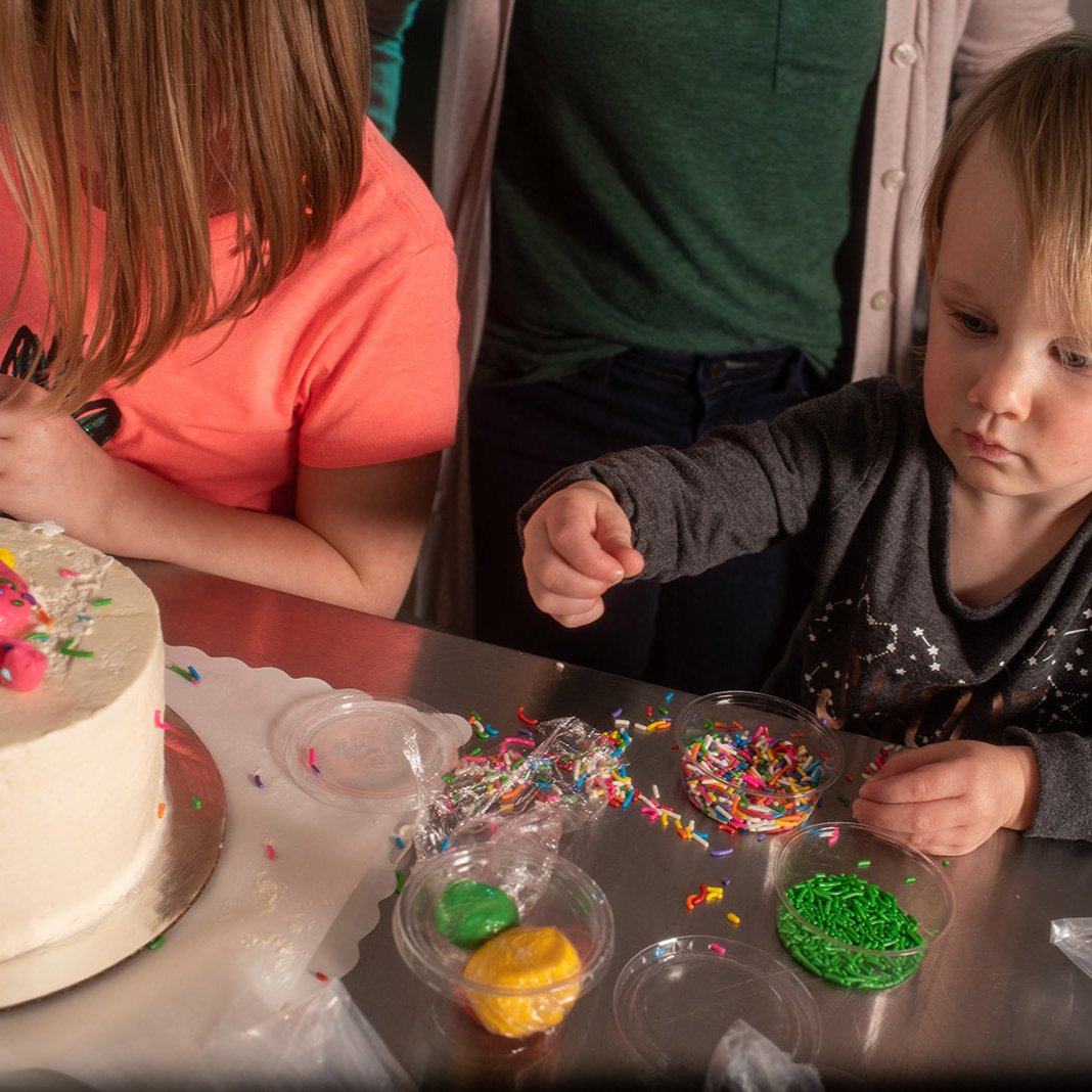 A little girl and a toddler are at a custom cake decorating party in the Portland area. They are decorating a white cake with pink frosting, fondant, and several colors of sprinkles. 