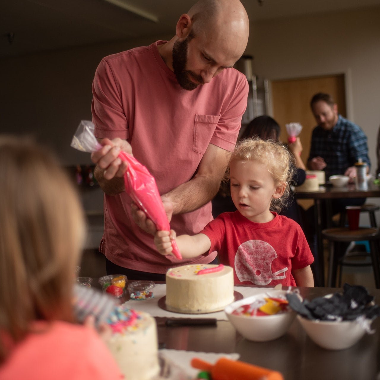 At a custom DIY cake decorating party in the Portland area, a father and son decorate a white cake with pink frosting from a piping bag. They are both concentrating and appear happy. Other groups of kids and parents are also decorating cakes. 