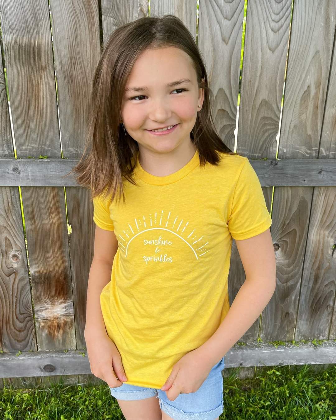 An eight-year-old girl wears the “sunshine & sprinkles” t-shirt. The color is vibrant and pops against her skin. The design in the center of the t-shirt spans horizontally almost all the way across her chest. 