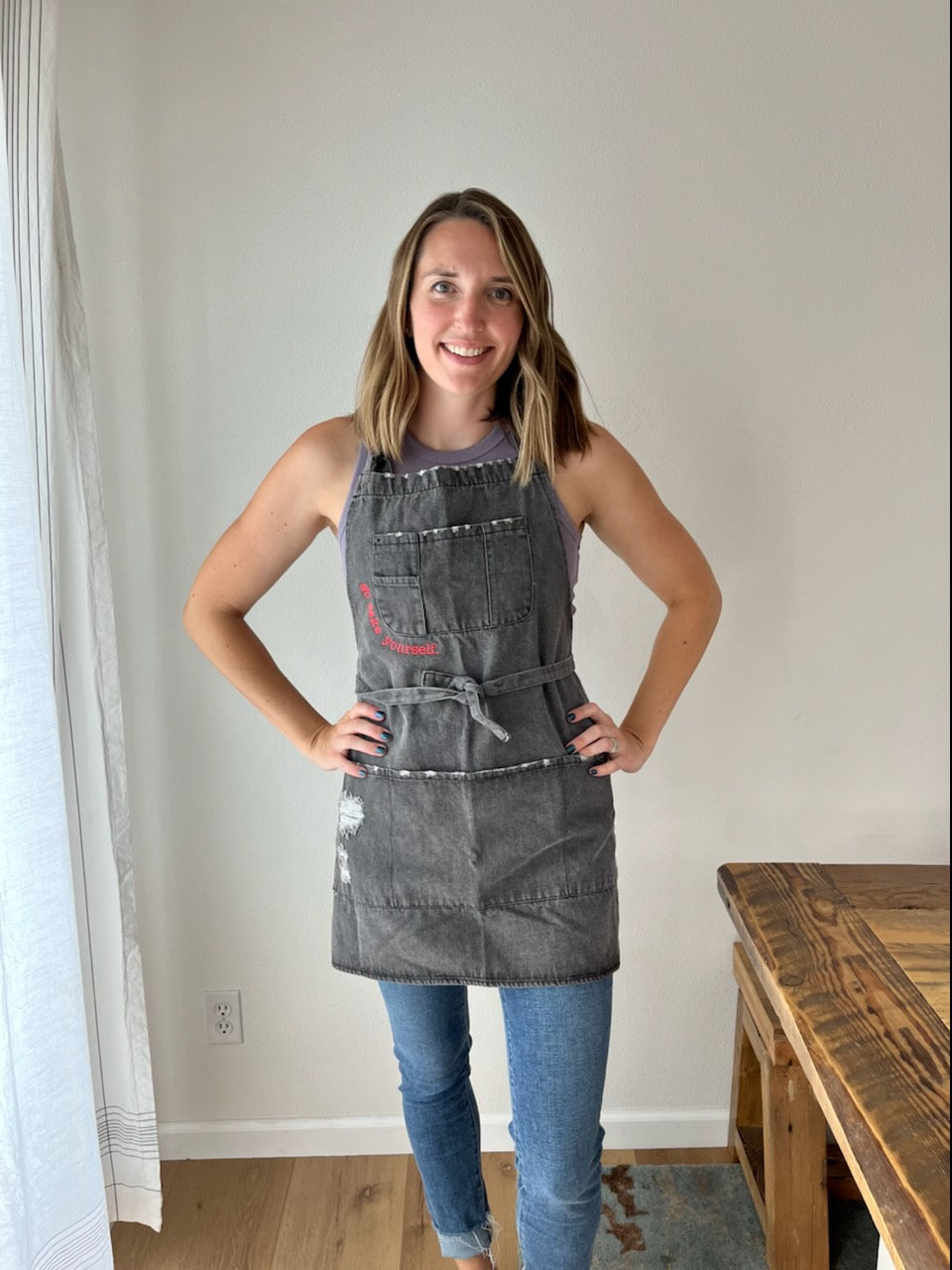 A woman standing 5 feet 9 inches is wearing the black distressed denim apron. The apron covers almost to her neck and extends down to mid-thigh. The straps loop around and are loosely tied in the front around her waist, slightly emphasizing her curves.