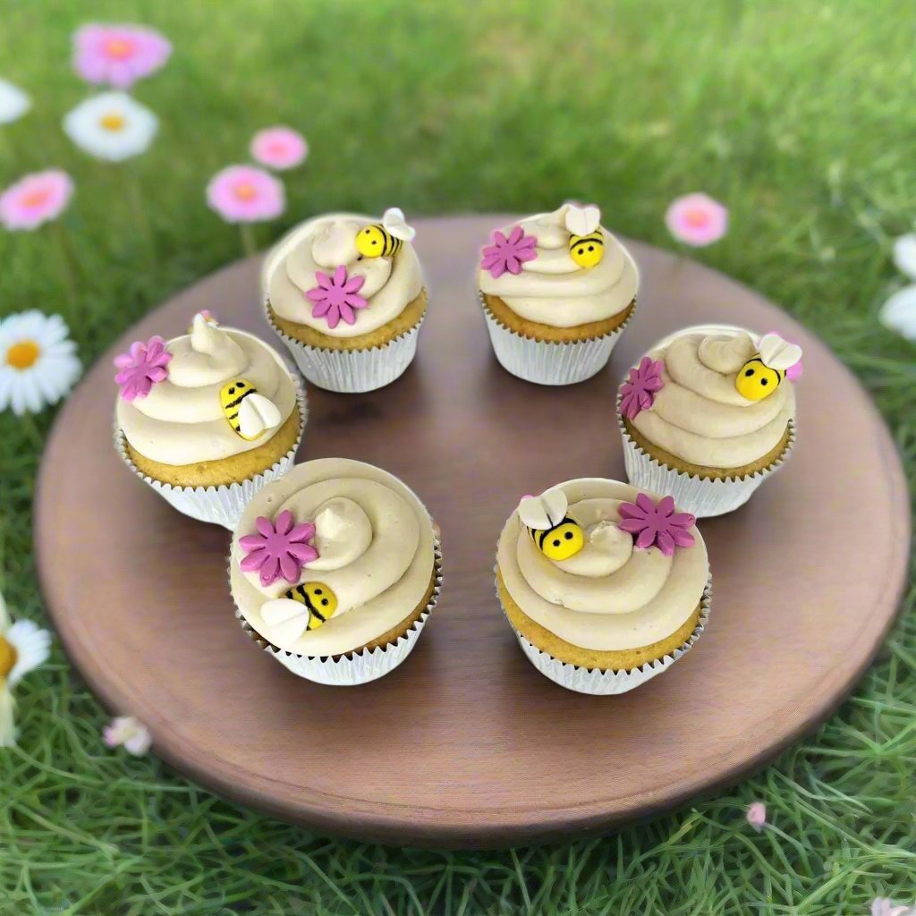 Finished bee-themed DIY cupcakes form a circle, each with a mound of white frosting on top that looks a honeycomb. Pink fondant flowers and yellow, pill-shaped fondant bees with white heart-shaped wings are on top of the frosting. Drawn in black edible marker are two dots for the bees' eyes and three black stripes along the bees' bodies.