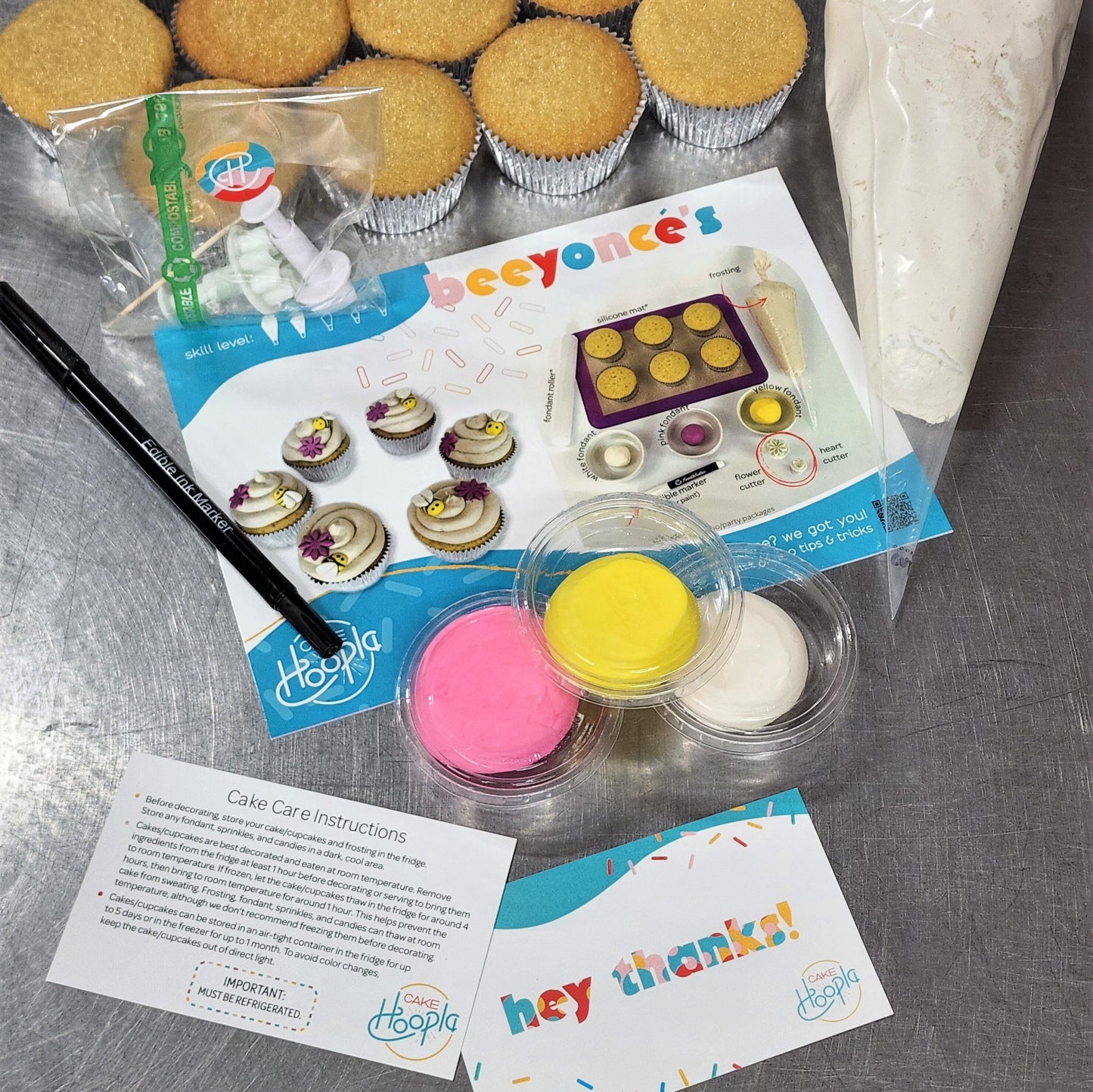 The ingredients and materials in a DIY Beeyonce's cupcake kit. 12 cupcakes, a large piping bag of white frosting, an edible black marker, pink, yellow, and white fondant, two fondant cutters in a compostable cellophane bag, a colorful instruction card with pictures, a cake care instruction card, and a thank you card.