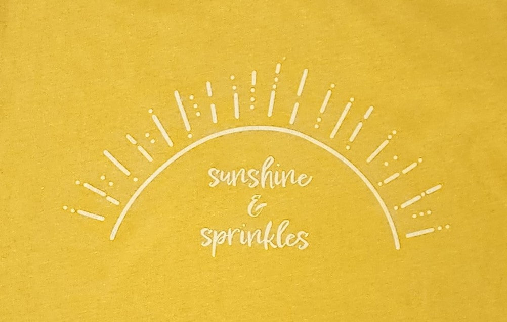 A closeup of the screen printing on the “sunshine & sprinkles t-shirt. “Sunshine & sprinkles” is written in a whimsical, almost cursive font across the center. The words are framed by an abstract half sun with rays of sprinkley sunshine shooting out. 