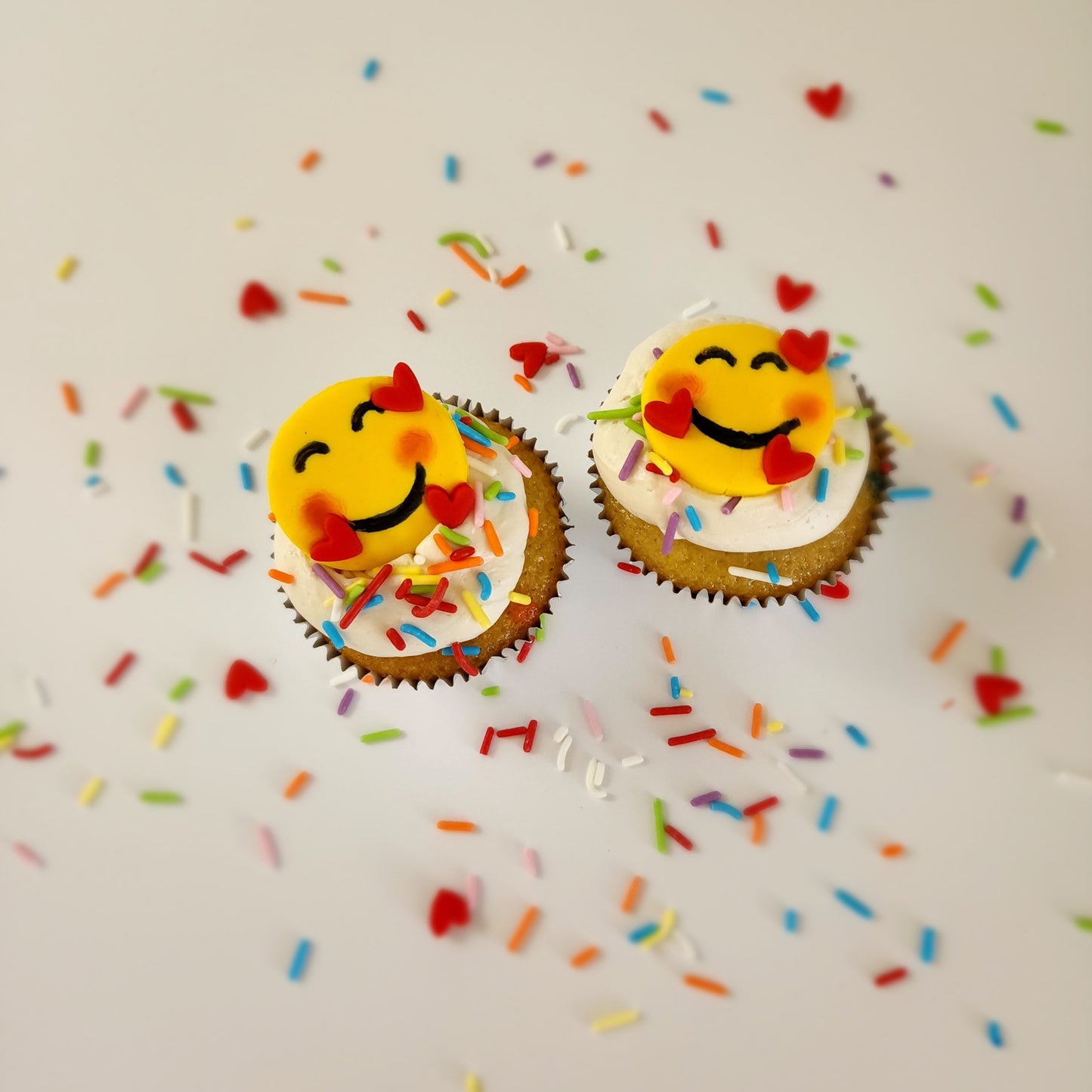 Emoji cupcakes frosted white with rainbow sprinkles and yellow smiling emoji faces with pink cheeks and three floating hearts; pre-decorated cupcakes available for shipping to the continental US.