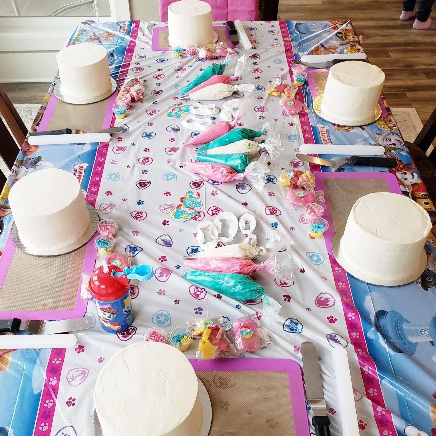 The setup for a custom dog-themed kids' cake decorating party the Portland area. A long table with bright Paw Patrol decorations contains white frosted cakes, piping bags with frosting, sprinkles, fondant, and tools for DIY cake decorating.